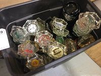 Photo showing a black plastic bin containing multiple clear glass ashtrays with various logos and inscriptions from hotels and businesses, along with one plastic bin without lid.