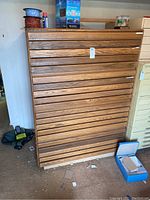 Front view of wooden blueprint cabinet showing four wide drawers with wood slat fronts and horizontal handles, medium-brown wood grain finish, placed on carpeted floor with miscellaneous items around.