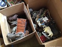 Two cardboard boxes containing a mix of buffalo figures made of plastic, resin and metal, plus one wooden or faux wood box.
