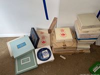 Photo showing stacks of boxed collectible plates including various Avon Christmas plates, Royal Copenhagen porcelain, and Norman Rockwell plates. Boxes vary in size and condition, some labels visible.