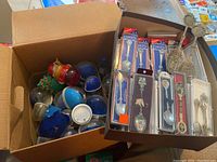 A box filled with multiple plastic snow globes of various colors including red, blue, and clear plastic, some missing internal water. A tray with multiple collectible souvenir spoons, some in original packaging, others loose with ornate designs and embossed travel landmarks.