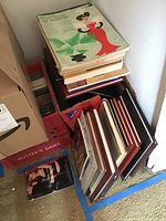 Stack of assorted commemorative yearbooks and music sheet books placed on floor with visible artwork on top book and a cardboard box nearby.