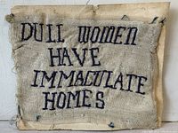 Front view of the cross stitch textile featuring the text 'DULL WOMEN HAVE IMMACULATE HOMES' on a worn fabric with frayed edges.