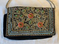 Front and back view of an antique rectangular clutch purse with detailed beadwork showing floral and leaf patterns, featuring green and purple stones and reflective beads on black fabric background. Long fabric strap visible.