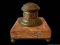 Side view showing square pink marble base with natural veins and four round metal feet, topped with ornate metal inkwell with floral and scroll embossed designs.