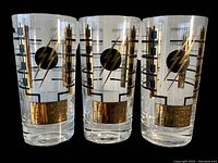 Three clear glass highball glasses decorated with retro atomic style black and gold geometric patterns and lightning bolt designs on a black background.
