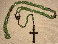 Full rosary laid out showing green glass beads, metal chain links, medallion and crucifix pendant on a beige fabric background.