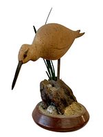 Front angle view of the hand carved shorebird decoy showing the bird leaning forward with a single eye visible, mounted on a detailed base with sand and grass blades on a round wooden platform.