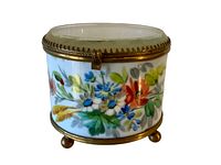 Oval hand painted glass canister with colorful floral design, brass hinged lid with toothed edge, and brass ball feet, shown from front angle