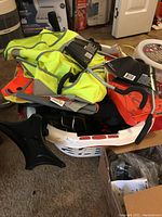 Assortment of reflective safety vests, bike seats, pedals, a white plastic basket containing additional bike gear and locks, and a black fanny pack.