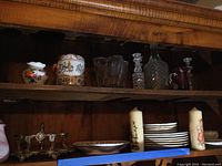Shelf with various decorative vases, crystal and glass decanters, two candles, and stacked plates and bowls below.