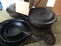 Photo showing three cast iron skillets stacked inside each other and two large lidded cast iron pots beside them, cardboard box on top.