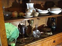 Wide shot of wooden cabinet shelves with porcelain dishes, silver plate items, vases, glass bowl, and paperweight.