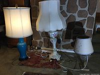 Three table lamps side by side on floor in front of stone wall, including blue ceramic, ornate floral metal, and clear glass base lamps.