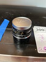 Front view with Leica-branded metal cap attached, lens resting on black surface alongside MaxSold notes card.