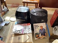 NuWave Power Air Fryer Oven with recipe book on the floor, adjacent to a cardboard box containing multiple stainless steel turkey kit accessories and the black NuWave Infrared Oven.
