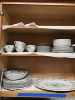 Photo shows several stacks of the dinnerware including cups, bowls, plates arranged on shelves inside a cabinet with Provencal floral pattern visible.