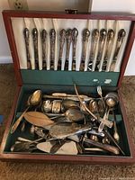 Full view of open wooden storage box with cream and green fabric lining holding silver plated flatware and assorted serving pieces.