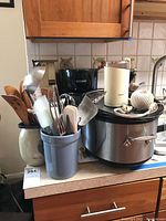 All kitchen appliances and utensils arranged on a counter including crockpot, coffee grinder, utensil holders, and coffee maker.