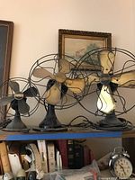Three vintage fans on a shelf: one black Peerless fan and two brass-blade fans.