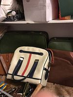 Photo showing white handbag with black handles and a vertical red stripe, green suitcases and brown large bag in background