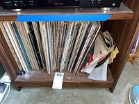 Photo showing a collection of vinyl records stored upright on a wooden shelf below an audio component, with visible wear and some damaged covers.