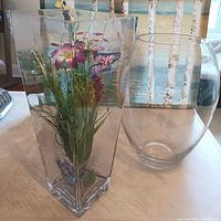 Photo showing two clear thick glass vases, one square with artificial flower arrangement and one oval-shaped clear vase.