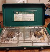 Top view of the 2 burner propane stove showing burners, grates, and instruction label inside the lid.