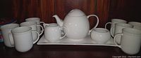 Full tea set arranged including teapot, milk pot, sugar pot, sugar pot lid with spoon, seven cups, and serving tray on a wooden surface