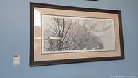 Framed artwork showing a moose behind tree branches, hung on a blue wall with visible frame and matting.