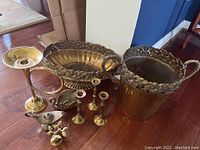 Full lot showing all brass items together on wood floor: large ice bucket, serving dish, candlesticks, genie lamp, swan decor.