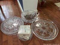Various crystal and glass serving pieces including trays and bowls arranged on wooden floor, some with intricate cut and etched designs.
