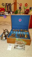 Wide shot showing open flatware chest on carpet with displayed gold-toned flatware, two metal teapots to left, and a clear plastic bag containing utensils in front.