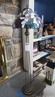 Full view of the floor lamp with stained glass shade, showing the metal base and overall height near bookshelf and stone wall.
