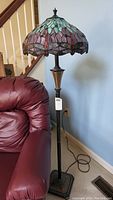 Full view of floor lamp with stained glass shade and dark metal post, next to burgundy chair and stair rail in background.
