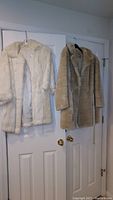Two fur coats hanging on door, beige beaver fur coat and white rabbit fur coat.