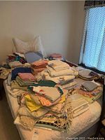 Various linens piled on bed including bedsheets, towels, quilts, placemats, and other fabric items in many colors and patterns