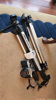 Two adjustable camera tripods laid horizontally on floor with a vintage Pentax camera nestled between them.