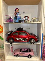 Overall lot view on three shelves: top shelf with dolls, scooters, toy dogs, and robot toy; middle shelf with red Volkswagen remote control car; bottom shelf with pink Mustang remote control car and remote control unit.