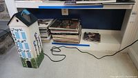 Photo shows a shelf with stacks of vinyl records, a pile of CDs, and a painted wooden birdhouse shaped item standing on the floor