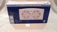 Boxed 9 inch dual fan window fan with thermostat shown from front.