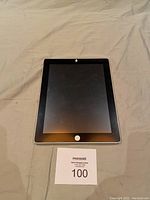 Front view of Apple iPad with black screen and white home button, displayed on a surface with MaxSold auction card.