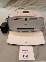 Front view of white HP Photosmart A516 photo printer showing card slots, front paper tray extended, power adapter adjacent