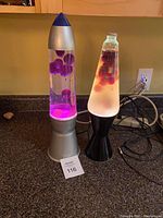 Two lava lamps on a counter: left lamp is rocket style with purple wax and silver base; right lamp is classic shape with black base, cloudy water, and purple wax.