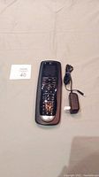 Photo showing Logitech Harmony One universal remote and charger on beige surface with lot number card.