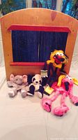 Wooden puppet theatre with four plush hand puppets arranged in front on a table; theatre has red accent and blue curtains.