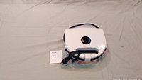 Top view of the white Magic Chef sandwich maker press with black handle and folded power cord around handle, placed on a gray surface with lot identification card beside it.