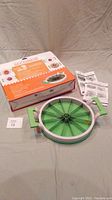 Green and white circular watermelon slicer with blade segments and handles, accompanied by a box and instructions on a gray surface.