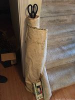 The full length of the cream colored fabric bolt with green floral pattern, showing it rolled up and standing upright at the base of a carpeted stairway with large black handled scissors inserted through the center.
