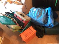 Photo showing green plastic tote with lid, red and black Plano tackle box, blue tarps, orange container, walking sticks, and sleeping bags gathered.